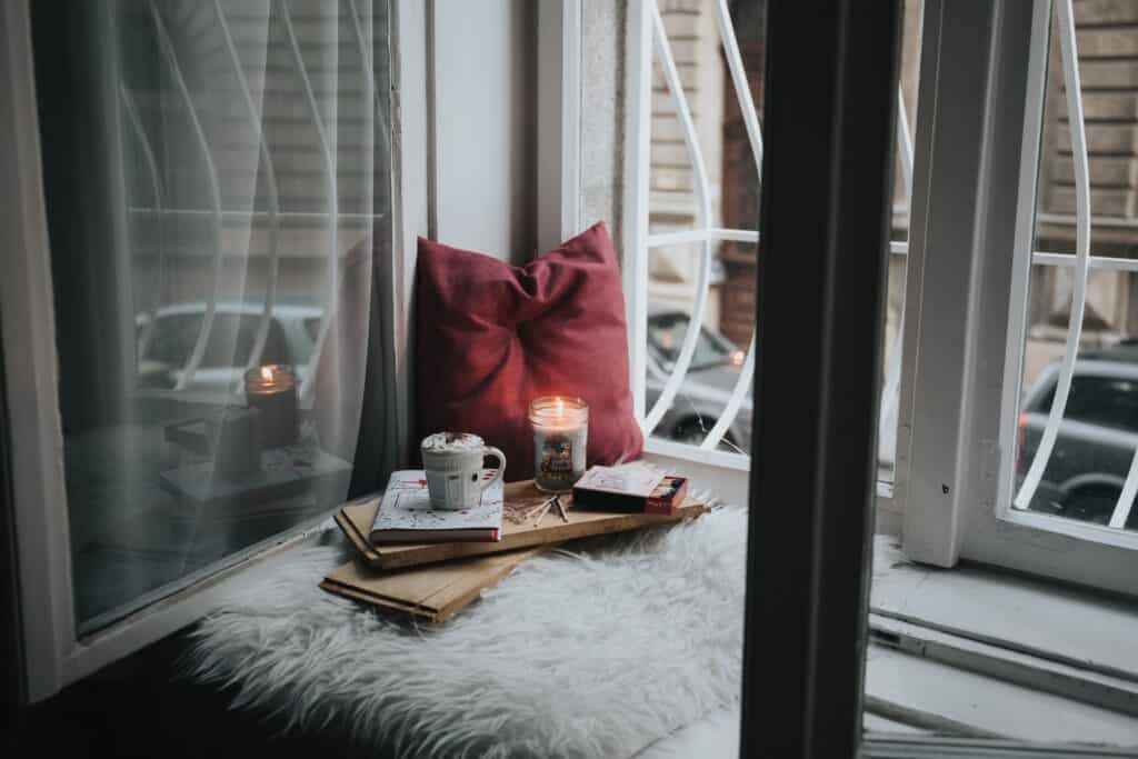 An enchanting cozy reading nook, with a comfy sitting arrangement nestled by a window, adorned with a soft blanket and candle and surrounded by stacks of books.